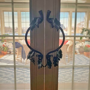 May include: A pair of black metal door handles with a vine design. The handles are attached to a white door with a window behind them.