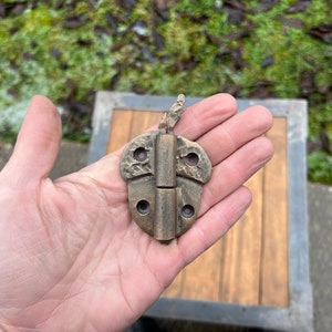 Puede incluir: Una bisagra de mariposa de latón con una superficie texturizada y dos juegos de agujeros para tornillos. La bisagra está cerrada y parece ser antigua.