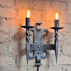 Puede incluir: Aplique de pared de hierro forjado con dos candelabros. El aplique tiene un diseño decorativo de metal martillado con brazos retorcidos y un soporte central. Dos bombillas encendidas en forma de llama emiten un brillo cálido.