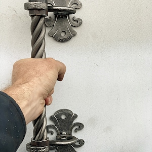 Puede incluir: Una manija de metal retorcido con un pomo decorativo en forma de bola y un pestillo a juego. La manija está unida a una pared blanca.