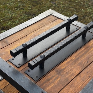 May include: Two black metal door handles with a textured finish are mounted on a wooden surface. The handles are rectangular with rounded edges and have a small knob on each end.