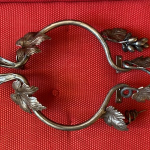 以下が含まれることがあります： つる植物と葉のデザインが施された、装飾的な金属製のドアノブのペア。ノブは暗い金属製で、素朴な仕上げが施されています。