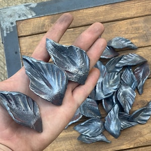 May include: A collection of metal leaf-shaped embellishments, with a hand holding a few of the pieces. The leaves are a silvery gray color and have a textured surface.