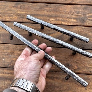 Może przedstawiać: Trzy rustykalne, młotkowane metalowe uchwyty do szuflad z chropowatym, fakturowanym wykończeniem. Uchwyty są srebrne i ułożone są w sposób przesunięty.