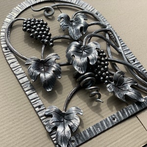 Puede incluir: Arte de pared decorativo de metal con un diseño de vid. La obra de arte incluye racimos de uvas, hojas detalladas y zarcillos rizados dentro de un marco arqueado. El metal tiene un acabado plateado.