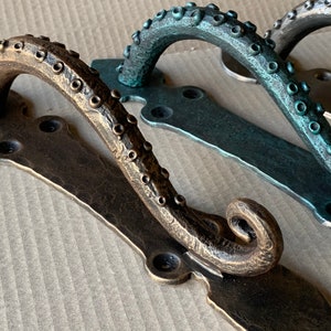 May include: Three octopus-shaped door handles in different finishes: bronze, teal, and a darker green. The handles are made of metal and have a detailed design.