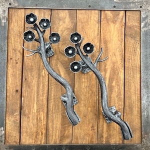 以下が含まれることがあります： 黒い金属の花がついた、スタイリッシュな枝の形をした装飾的な金属製のドアノブ2つ。ノブは、濃い茶色のステインが塗られた木製ドアに取り付けられています。