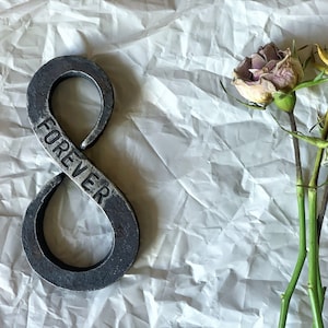 May include: A metal infinity symbol with the word "FOREVER" engraved on it. The symbol is on a white, crumpled background with three dried roses.