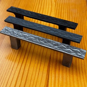 Puede incluir: Tres tiradores de armario en diferentes acabados. El tirador superior es negro, el tirador del medio es negro y el tirador inferior es plateado. Los tres tiradores tienen un acabado texturizado martillado.