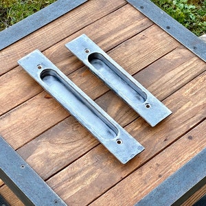 Könnte beinhalten: Zwei silberne Metall-Schubladengriffe in rechteckiger Form mit einem eingelassenen Griff. Die Griffe befinden sich auf einer Holzoberfläche mit einer dunkelbraunen Beize.