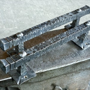 May include: A hand-forged iron door handle with a hammered finish. The handle has a square shape and is attached to a metal plate.