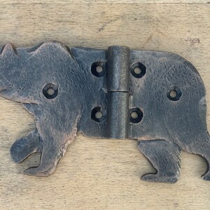 May include: A decorative, black metal hinge in the shape of a bear. The hinge has a rustic, textured finish and features multiple screw holes for mounting. The bear is depicted in a walking pose, adding a whimsical touch.