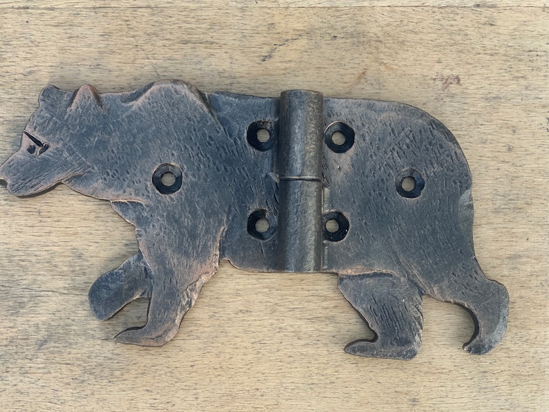 May include: A decorative, black metal hinge in the shape of a bear. The hinge has a rustic, textured finish and features multiple screw holes for mounting. The bear is depicted in a walking pose, adding a whimsical touch.