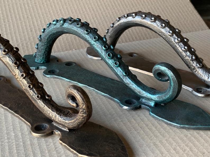 May include: Three octopus-shaped hooks with a textured surface. The hooks are made of metal and have a dark green, bronze, and silver finish. The hooks are mounted on a flat metal plate.
