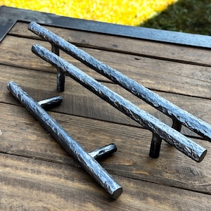 Puede incluir: Dos tiradores de cajón de metal martillado, uno largo y otro corto, con un acabado rústico. Los tiradores se muestran sobre una superficie de madera.