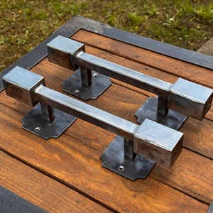Puede incluir: Dos tiradores de puerta metálicos con diseño cuadrado. Los tiradores son plateados y están sujetos a una base de metal oscuro. Los tiradores están montados sobre una superficie de madera con un marco de metal oscuro.