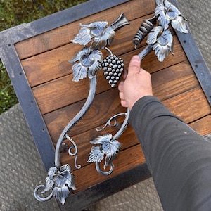 May include: Hand-forged metal grape vine decor with leaves and a cluster of grapes. The metalwork is silver-colored and detailed, with a rustic, handcrafted appearance. The vine is displayed on a wooden surface.
