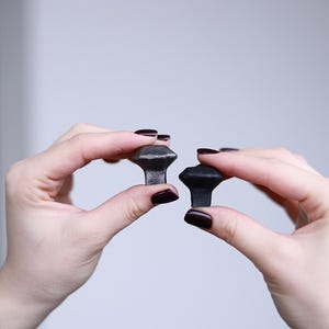 Könnte beinhalten: Zwei schwarze Metall-Schubladengriffe mit einem rautenförmigen Design. Die Griffe werden in den Händen einer Person gehalten.