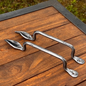 Può includere: Due maniglie per cassetti argentate a forma di foglia. Le maniglie sono curve con un design a foglia a un'estremità e una base piatta e forata all'altra. Poggiano su una superficie di legno con una cornice in metallo scuro.