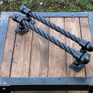 May include: Two black, twisted metal handles with ball finials are mounted on a wooden surface. The wood has a natural grain and is framed by a dark metal border. The handles are attached to the wood with square metal plates.