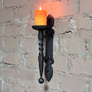 Puede incluir: Un aplique de pared de hierro forjado negro con una vela naranja encendida. El aplique tiene un tallo retorcido y una placa trasera decorativa, montado en una pared de ladrillo de color claro. La llama de la vela brilla intensamente.