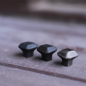 May include: Three square-shaped cabinet knobs in black and silver finishes. The knobs are arranged in a row on a wooden surface.