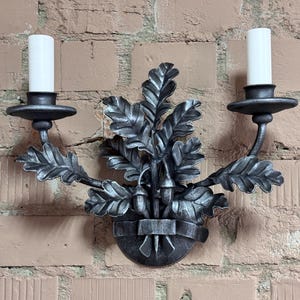 May include: A decorative, two-light wall sconce made of dark metal, featuring stylized oak leaves and acorns. The sconce holds two white candle-shaped lights. The sconce is mounted on a brick wall.