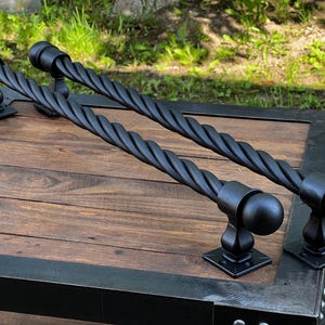 Pode incluir: Duas puxadores de gaveta de metal preto com um design de corda torcida e terminais redondos. As puxadores são montados em uma superfície de madeira.