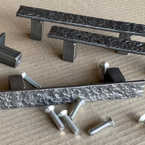 May include: Five metal drawer pulls with a hammered finish. The pulls are in various sizes and shapes, including a long rectangular pull, a shorter rectangular pull, and a curved pull. The pulls are shown on a brown surface with several screws.