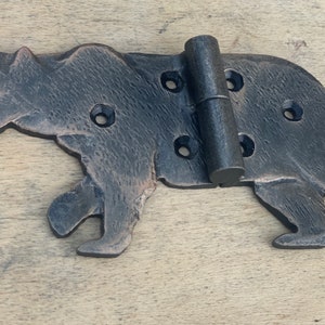 May include: A decorative, cast iron hinge in the shape of a bear. The hinge is dark brown with visible texture and features multiple screw holes. The bear is depicted in a walking pose, with the hinge mechanism in the center.