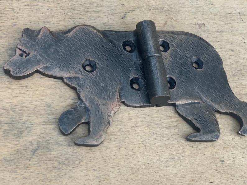 May include: A decorative, cast iron hinge in the shape of a bear. The hinge is dark brown with visible texture and features multiple screw holes. The bear is depicted in a walking pose, with the hinge mechanism in the center.