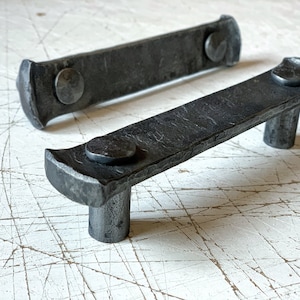 May include: Two black metal drawer pulls with rounded edges and a hammered finish. The pulls have a single, round, protruding knob on each end.