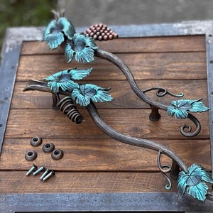 May include: Two ornate metal door handles shaped like grapevines with leaves and a cluster of grapes. The handles are on a wooden surface with screws and metal plates.