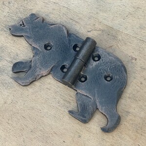 May include: A decorative, black metal hinge in the shape of a bear. The hinge has a rustic, textured finish and includes several pre-drilled holes for mounting. The hinge is designed to be used on a door or cabinet.