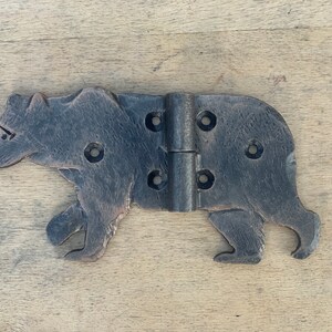 May include: A rustic, dark metal hinge in the shape of a bear. The hinge has a detailed bear silhouette with visible screw holes for mounting. The metal has a textured, aged appearance, suggesting a vintage or handcrafted item.