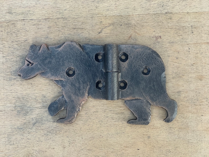 May include: A rustic, dark metal hinge in the shape of a bear. The hinge has a detailed bear silhouette with visible screw holes for mounting. The metal has a textured, aged appearance, suggesting a vintage or handcrafted item.
