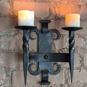 May include: A black wrought iron wall sconce with two candle holders, each holding a white candle. The sconce is mounted on a brick wall. The sconce has a decorative design with curved elements and a central mounting plate.