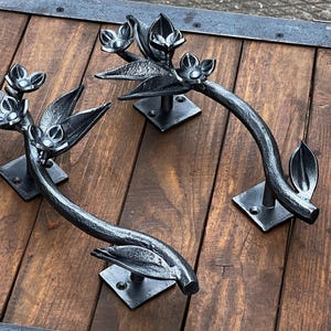 May include: Two decorative, wrought iron door handles with a floral design. The handles are dark gray and feature stylized leaves and flowers. They are mounted on square plates and set against a wooden surface.