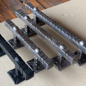May include: Set of four handcrafted metal door pulls. The pulls have a textured, hammered appearance with square bases and decorative rivets. Two are black, and two are a dark gray color. The hardware is designed for a rustic or industrial aesthetic.