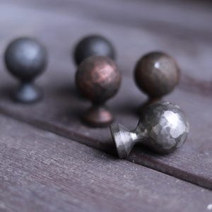 Könnte beinhalten: Fünf verschiedene Metall-Schrankknöpfe mit gehämmerter Oberfläche. Die Knöpfe sind in verschiedenen Grautönen und Kupferfarben.