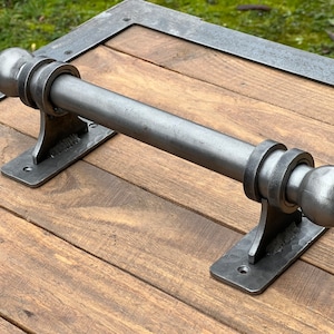 Puede incluir: Una manija de puerta de metal plateado con extremos redondeados y una barra cilíndrica. La manija está montada en una superficie de madera.