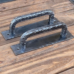 Może przedstawiać: Dwa czarne metalowe uchwyty do drzwi z młotkowanym wykończeniem. Uchwyty są zamontowane na prostokątnych płytach.