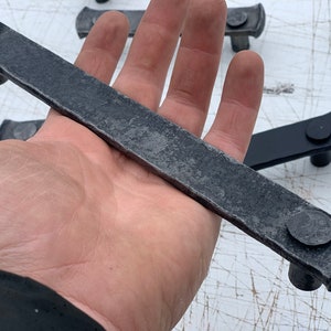 May include: A long, rectangular, black metal drawer pull with rounded edges and a hammered finish. The pull has two round, metal knobs on either end.