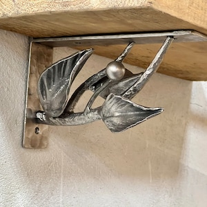 May include: A silver metal shelf bracket with a leaf design. The bracket is attached to a wooden shelf.