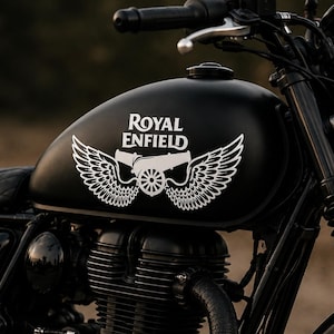 May include: A matte black Royal Enfield motorcycle featuring a white graphic on the fuel tank that displays the text Royal Enfield above a cannon flanked by two large wings. The motorcycle has a quilted seat, a round headlight, and black engine.
