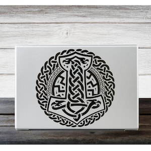 Puede incluir: Un portátil plateado con un diseño de martillo Mjolnir vikingo negro. El martillo está rodeado por un patrón de nudos entrelazados circular. El diseño está centrado en la tapa del portátil, creando un contraste llamativo.