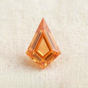 Könnte beinhalten: Ein drachenförmiger Edelstein mit leuchtend orangefarbenem Ton und facettierter Oberfläche. Der Stein hat eine spitze Ober- und Unterseite mit abgewinkelten Kanten und zeigt interne Reflexionen sowie geometrische Muster durch den Schliff.