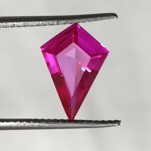 May include: A kite-shaped, faceted gemstone in a vibrant, saturated hot pink colour. The stone features a symmetrical geometric cut with multiple triangular facets that reflect light, held securely between the tips of two metal tweezers.