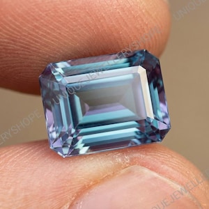 Puede incluir: Una gema rectangular facetada con una mezcla de tonos azules, morados y turquesas. La gema se sostiene entre dos dedos, mostrando sus superficies pulidas y variaciones internas de color. Las facetas reflejan la luz, creando un efecto brillante.