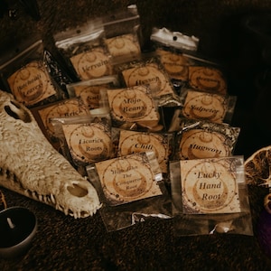 Op de afbeelding: Een assortiment kleine, doorzichtige plastic zakjes gevuld met kruiden en wortels, elk gelabeld met een handschrift. De labels bevatten namen als "Patchouli Leaves", "Mojo Beans" en "Lucky Hand Root". Een krokodillenschedel en gedroogde sinaasappelschijfjes zijn ook zichtbaar.
